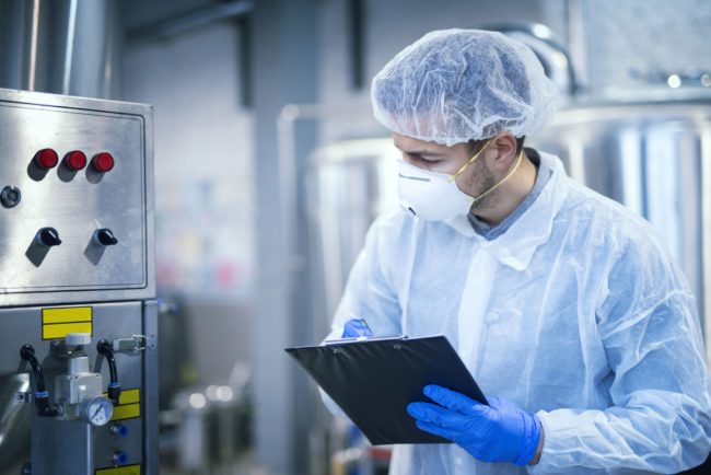 Technologist expert in protective uniform with hairnet and mask taking parameters from industrial machine in food production plant. food manufacturing